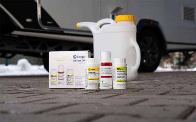 Drei kleine Fläschchen und eine Schachtel auf gepflastertem Boden, dahinter ein weißer Kanister mit gelbem Verschluss und ein Wohnwagen
