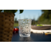 Helder, gestructureerd drinkglas op een donkere buitentafel; op de achtergrond onscherp een bord met groene bladeren, cherrytomaten en een plantenpot