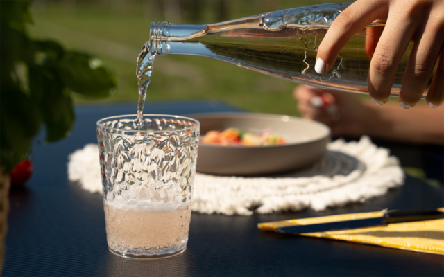 Hand giet lichtroze vloeistof uit een doorzichtige glazen fles in een gestructureerd drinkglas op een donkere tafel; op de achtergrond een kom op een gehaakte placemat, geel bestek en een vage plant