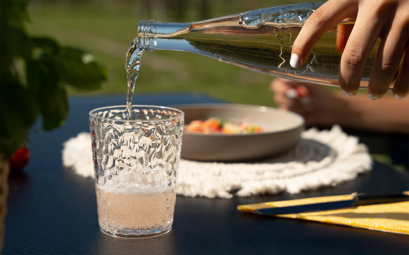 Hand giet lichtroze vloeistof uit een doorzichtige glazen fles in een gestructureerd drinkglas op een donkere tafel; op de achtergrond een kom op een gehaakte placemat, geel bestek en een vage plant
