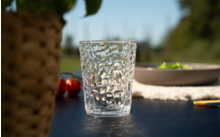 Verre transparent à surface texturée sur une table extérieure sombre ; en arrière-plan flou, une assiette avec des feuilles vertes, des tomates cerises et un pot de plante