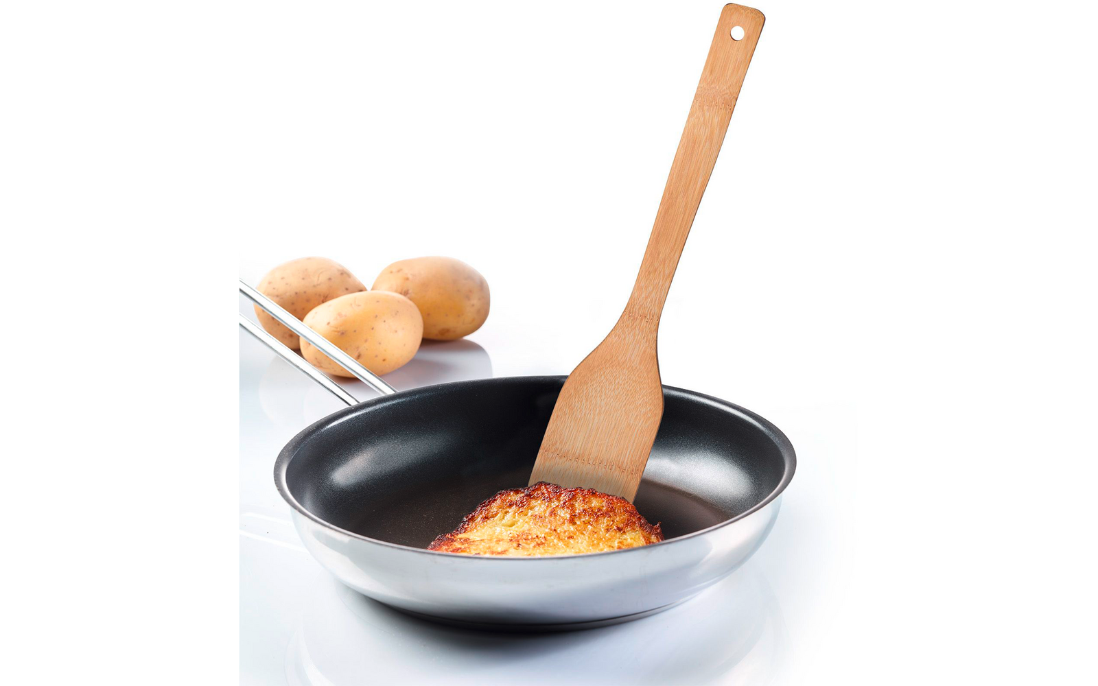 Stainless-steel frying pan with black nonstick interior holding a round golden-brown potato piece lifted by a wooden spatula; three raw potatoes in the background on a white surface