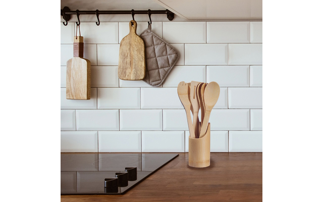 Kitchen countertop with cooktop, wooden utensils in a holder; two wooden cutting boards and a gray oven mitt hanging on a rail in front of white tiled backsplash