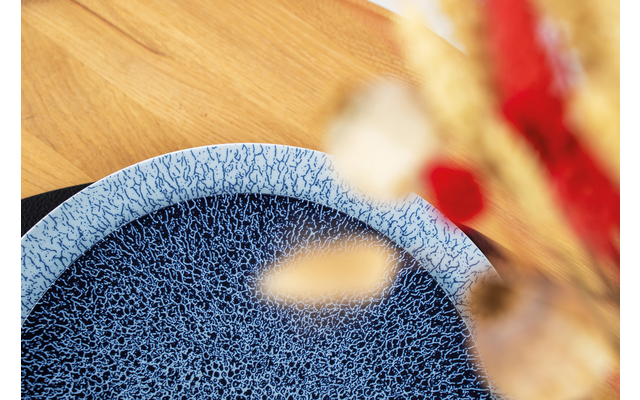 Nahaufnahme eines blauen, strukturierten Tellers auf einem Holztisch, rechts verschwommen Blumen