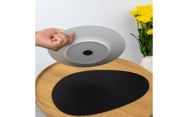 Hand hebt einen weißen Teller über einem runden Holztisch mit schwarzer ovaler Tischunterlage; rechts eine weiße Vase mit gelben Blumen