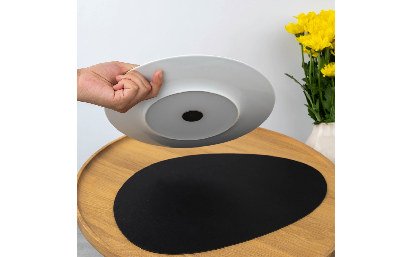 Hand hebt einen weißen Teller über einem runden Holztisch mit schwarzer ovaler Tischunterlage; rechts eine weiße Vase mit gelben Blumen