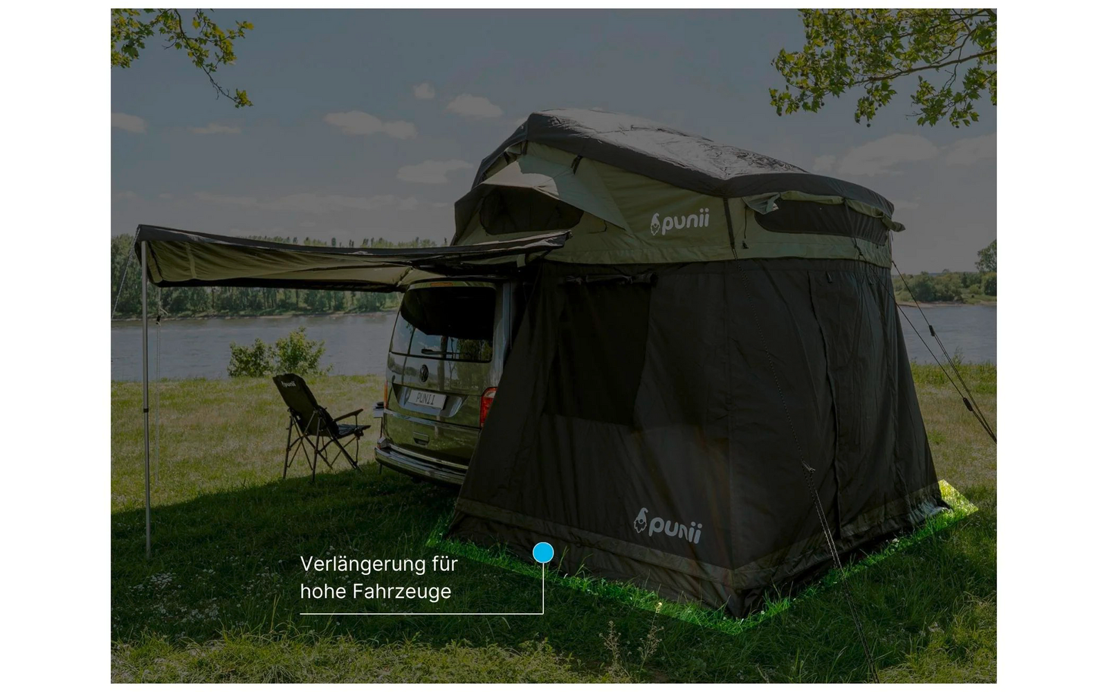 Van with roof-top tent and attached dark fabric annex, folding chair on the shore of a lake, trees and sky in the background