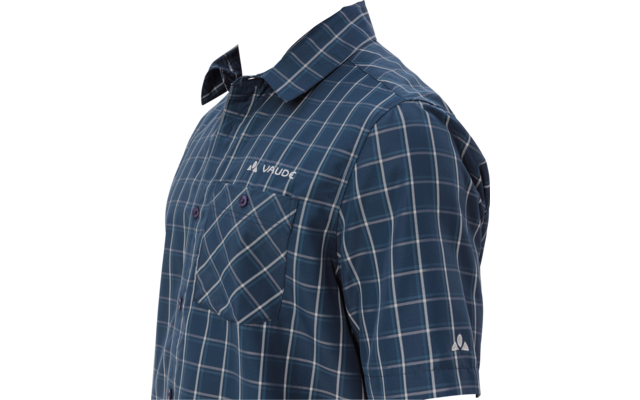 Person with short dark hair wearing a blue checkered short-sleeve shirt with chest pockets and white embroidery, looking to the left, white background