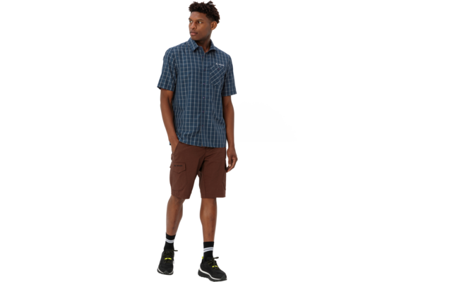 Mann in blau kariertem Kurzarmhemd und braunen Cargoshorts, mit schwarzen Sneakers und weißen Socken, steht vor weißem Hintergrund und blickt nach links