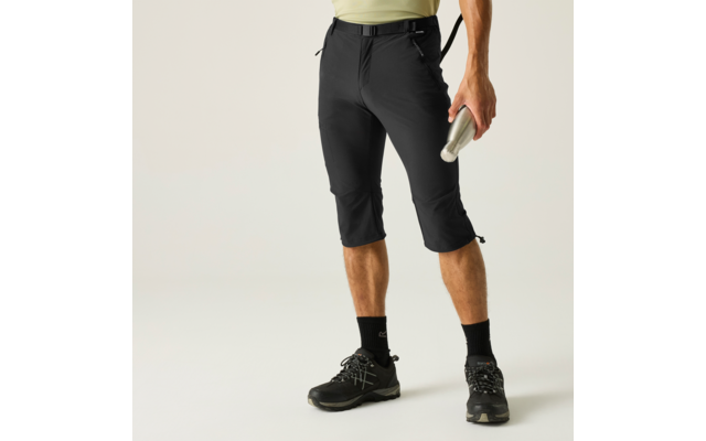 Unterkörper mit schwarzer dreiviertellanger Hose und schwarzem Gürtel, schwarzen Socken und Schnürschuhen; rechte Hand hält eine silberne Trinkflasche; hellgrünes Oberteil am Bund sichtbar