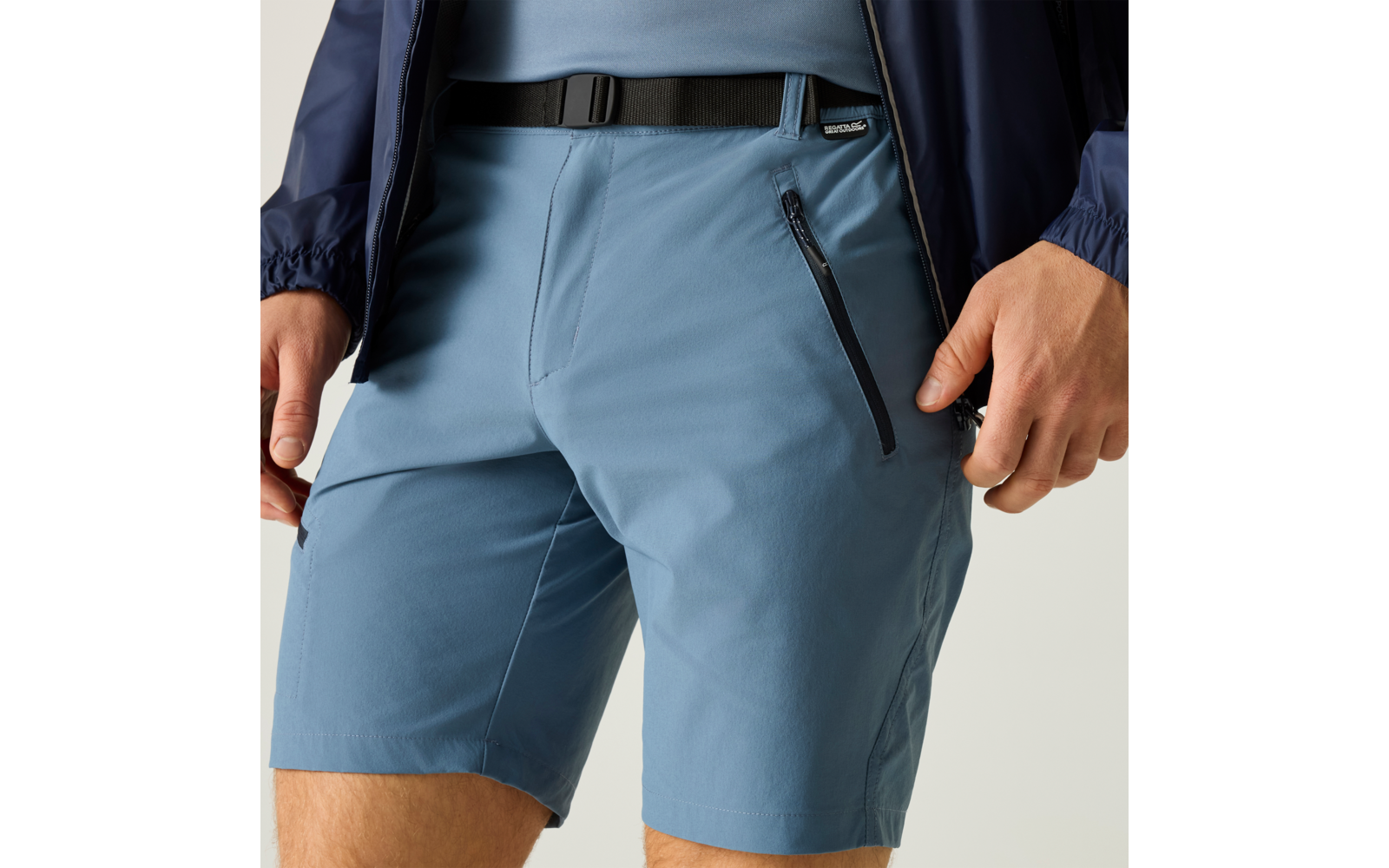 Unterkörper und Oberschenkel einer Person, hellblaue Shorts mit schwarzer Reißverschlusstasche, schwarzer Gürtel mit Schnalle, dunkelblauer Jackensaum, Hände seitlich sichtbar