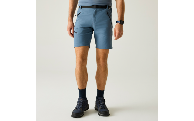 Unterkörper einer Person von der Hüfte bis zu den Füßen, blaue Outdoor-Shorts mit sichtbaren Reißverschlusstaschen und Gürtel, dunkle Socken, dunkle Wanderschuhe, Armbanduhr am linken Handgelenk, heller einfarbiger Hintergrund