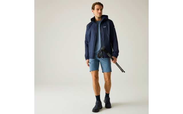 Mann in blauer Jacke, blauem T‑Shirt und Shorts, mit Wanderschuhen, hält ein zusammengeklapptes Stativ