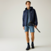 Mann in blauer Jacke, blauem T‑Shirt und Shorts, mit Wanderschuhen, hält ein zusammengeklapptes Stativ
