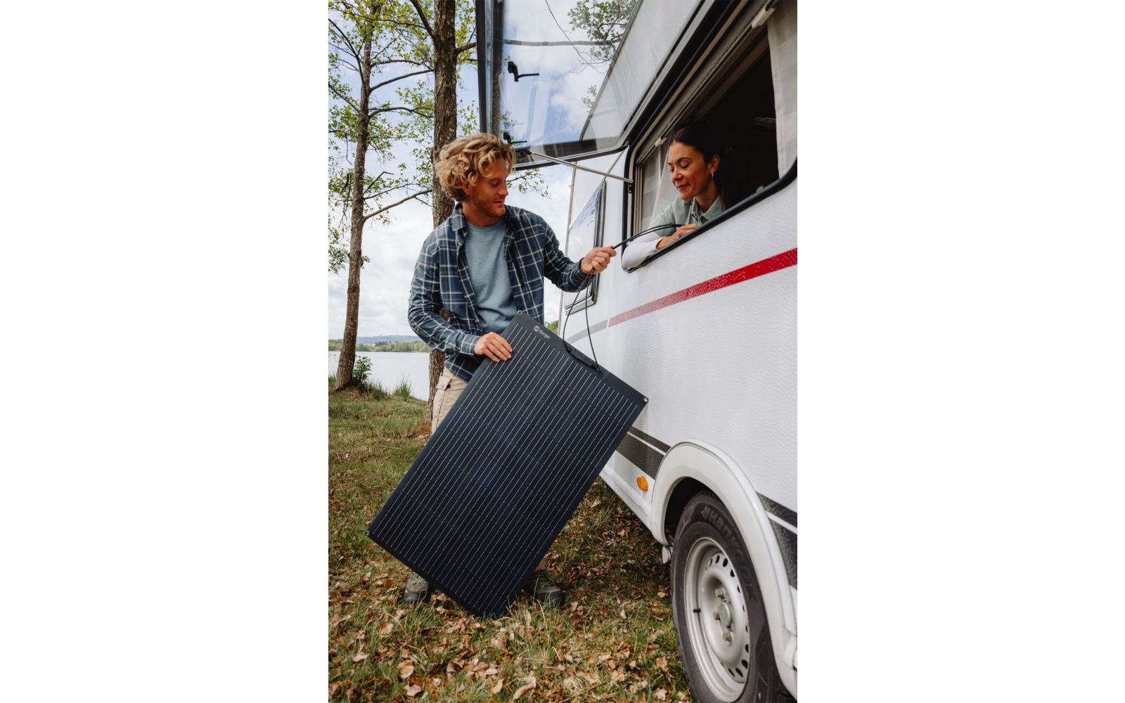 Un homme tient un grand panneau solaire et tend un câble à une femme qui sort la main par la fenêtre d'un camping-car blanc ; des arbres et un lac en arrière-plan
