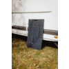 Un panneau solaire appuyé contre le flanc d'une caravane blanche sur de l'herbe avec des feuilles tombées; des câbles noirs reposent sur le panneau; la paroi de la caravane présente des bandes horizontales, un réflecteur orange et des ombres d'arbres