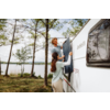 Un homme et une femme fixent une toile bleu foncé sur le côté d'un camping-car blanc. Un lac et des arbres en arrière-plan