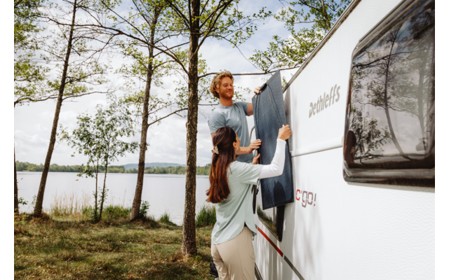 Un homme et une femme fixent une toile bleu foncé sur le côté d'un camping-car blanc. Un lac et des arbres en arrière-plan