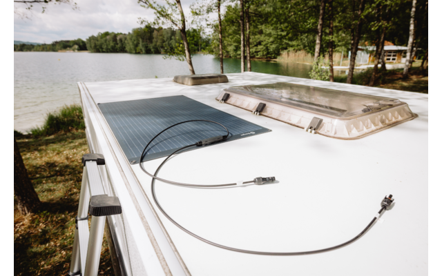 Toit de véhicule blanc avec panneau solaire, deux câbles détachés et un hublot de toit ; lac et arbres en arrière-plan