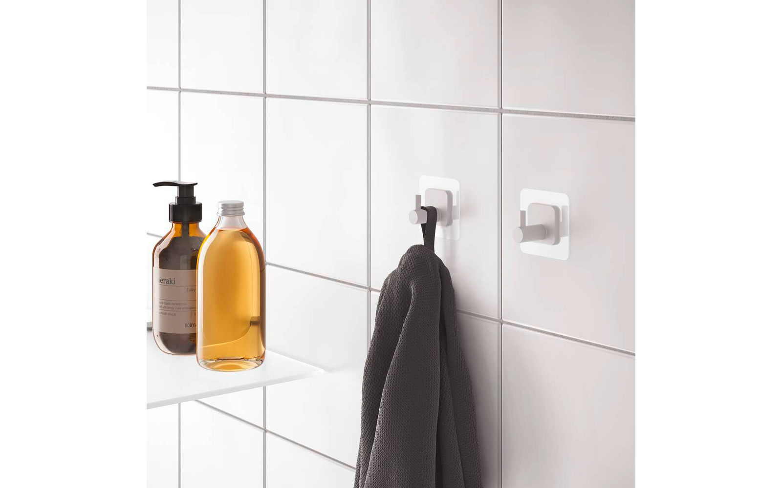 Weiße Fliesenwand mit zwei selbstklebenden Haken; an einem hängt ein dunkelgraues Handtuch; links auf einem weißen Regal zwei Flaschen, eine mit Pumpspender