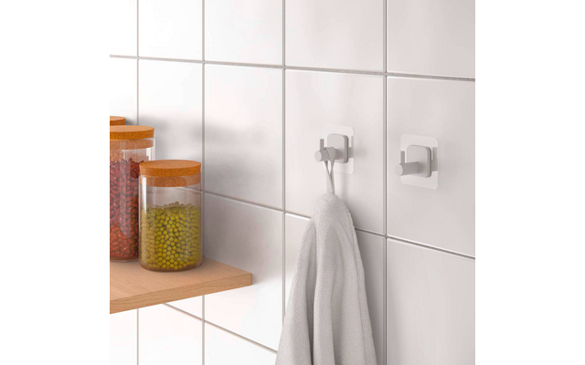 Weiß geflieste Wand mit zwei weißen Klebehaken, an einem hängt ein weißes Handtuch; links Holzregal mit drei Glasbehältern mit orangefarbenen Deckeln und rotem, grünem und gelbem Inhalt