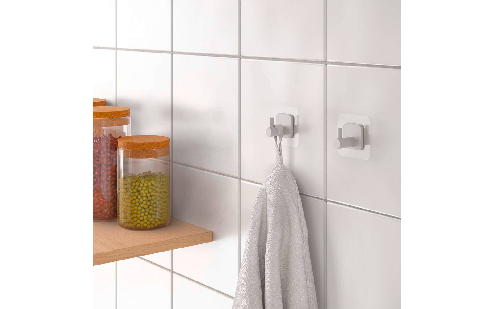 Weiß geflieste Wand mit zwei weißen Klebehaken, an einem hängt ein weißes Handtuch; links Holzregal mit drei Glasbehältern mit orangefarbenen Deckeln und rotem, grünem und gelbem Inhalt