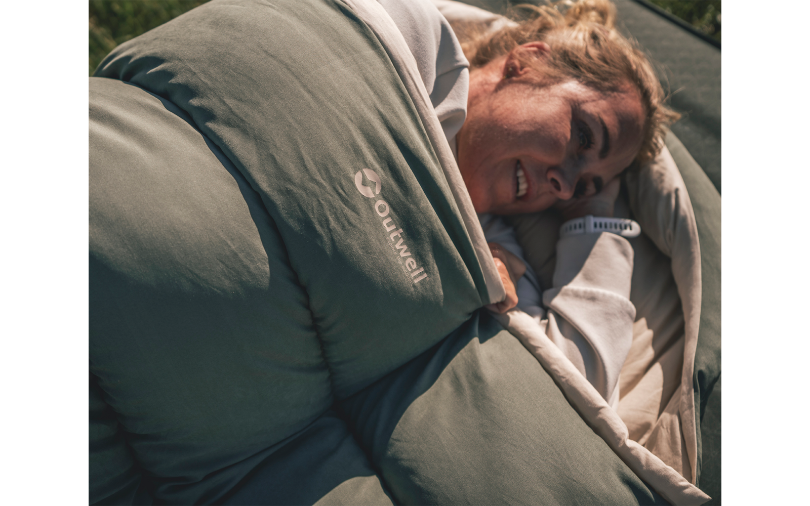 Persoon ligt in een groene slaapzak met zichtbaar Outwell-logo op een lichte ondergrond, gezicht deels zichtbaar, hand bij de opening van de slaapzak