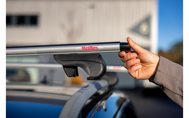 Hand mit Jackenärmel hält silberne Dachträger-Querstange mit rotem "MontBlanc"-Logo, montiert auf Autodach; unscharfes Gebäude und Himmel im Hintergrund