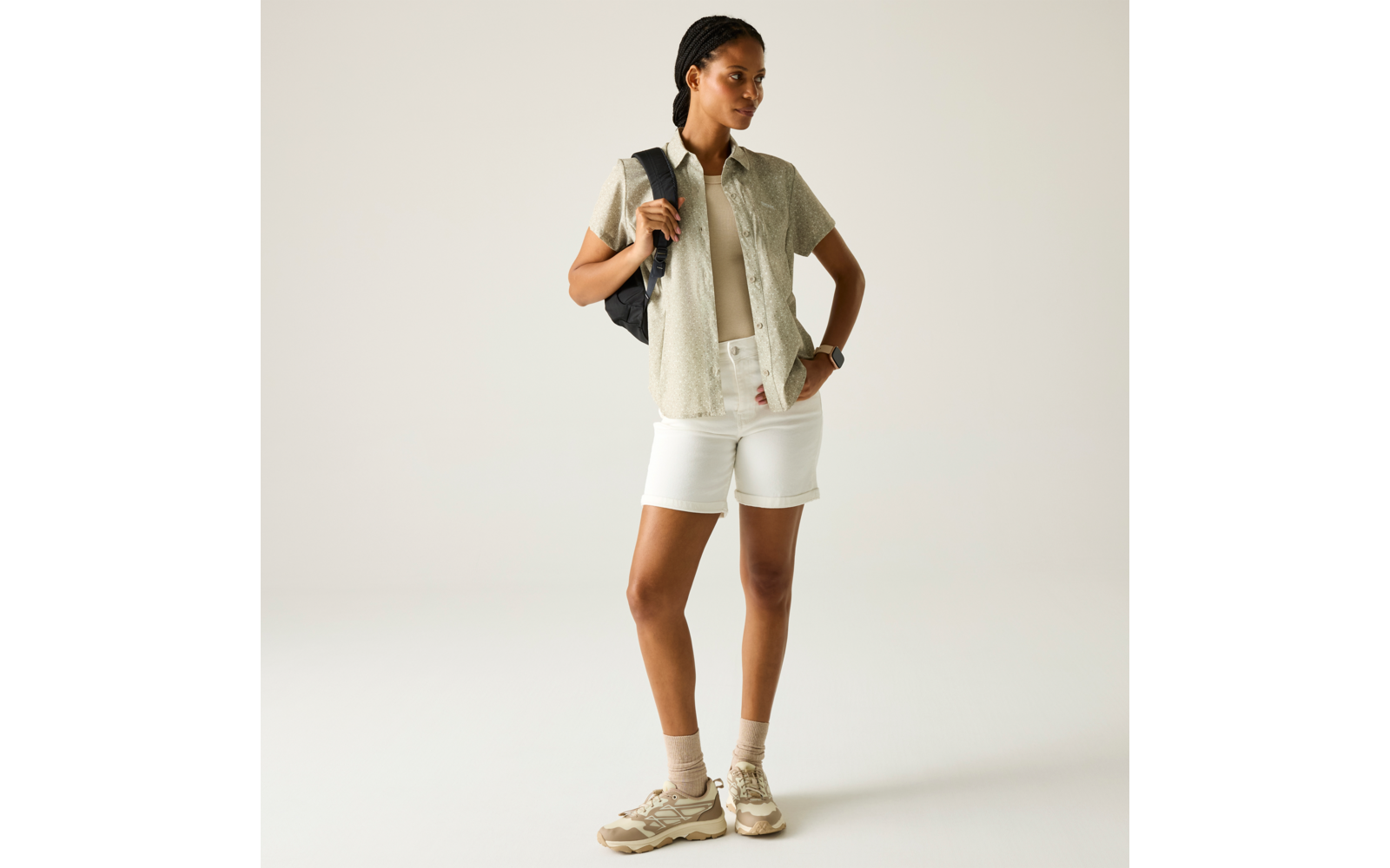 Person steht mit Rucksack über einer Schulter, trägt helles kurzärmeliges Hemd, beiges Top, weiße Shorts, beige Sneakers und Socken, eine Hand an der Hüfte, schaut nach rechts, heller einfarbiger Hintergrund