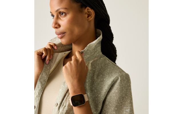 Person mit geflochtenen Haaren hält den Kragen einer Jacke mit kleinem Muster; trägt hellen Pullover und Smartwatch, neutraler Hintergrund