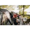 Zwei Personen an einem Campingplatz neben einem Zelt. Eine Person steht an einem tragbaren Grill, die andere sitzt an einem Tisch mit Getränken. Im Hintergrund Bäume, ein See und eine Hängematte