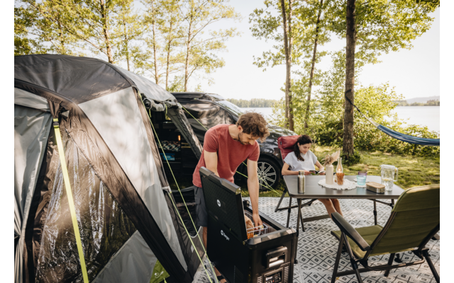 Zwei Personen an einem Campingplatz neben einem Zelt. Eine Person steht an einem tragbaren Grill, die andere sitzt an einem Tisch mit Getränken. Im Hintergrund Bäume, ein See und eine Hängematte