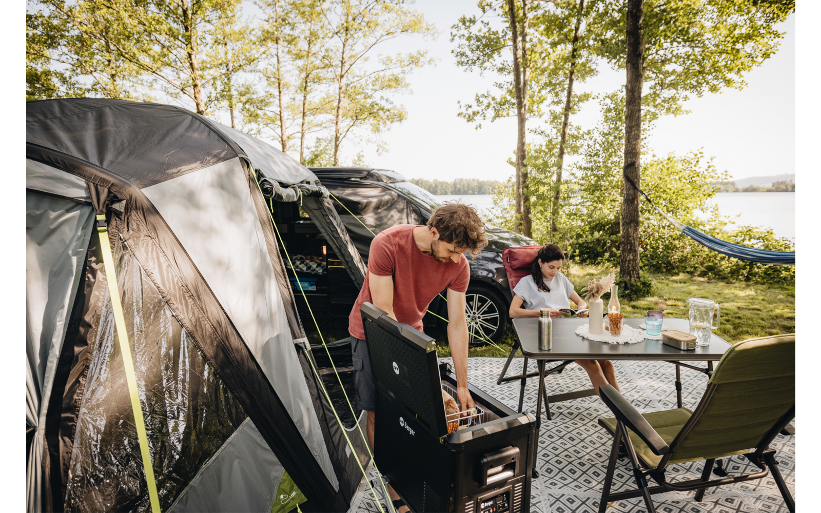 Zwei Personen an einem Campingplatz neben einem Zelt. Eine Person steht an einem tragbaren Grill, die andere sitzt an einem Tisch mit Getränken. Im Hintergrund Bäume, ein See und eine Hängematte