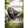 Person in rotem T‑Shirt steht neben offenem Campingvan und Ausrüstung; sitzende Person mit Kind auf dem Schoß an Campingtisch; See und bewaldetes Ufer im Hintergrund