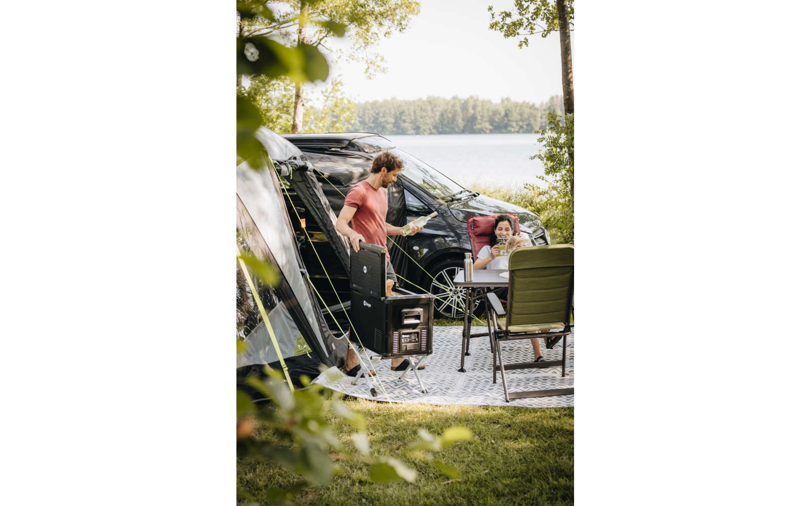 Person in rotem T‑Shirt steht neben offenem Campingvan und Ausrüstung; sitzende Person mit Kind auf dem Schoß an Campingtisch; See und bewaldetes Ufer im Hintergrund