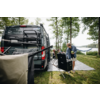 Grauer Campervan steht auf einer Wiese unter Bäumen neben einem See. Vor dem Van steht eine Person an einer geöffneten tragbaren Kochbox und hält etwas in den Händen. Zwei Personen sitzen auf Campingstühlen, eine Hängematte hängt zwischen Bäumen, ein