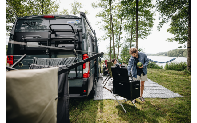 Grauer Campervan steht auf einer Wiese unter Bäumen neben einem See. Vor dem Van steht eine Person an einer geöffneten tragbaren Kochbox und hält etwas in den Händen. Zwei Personen sitzen auf Campingstühlen, eine Hängematte hängt zwischen Bäumen, ein