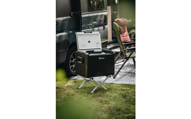 Offene schwarze tragbare Kühlbox auf Metallständer vor einem Van, daneben ein gepolsterter Campingstuhl und eine Bodenmatte auf Gras