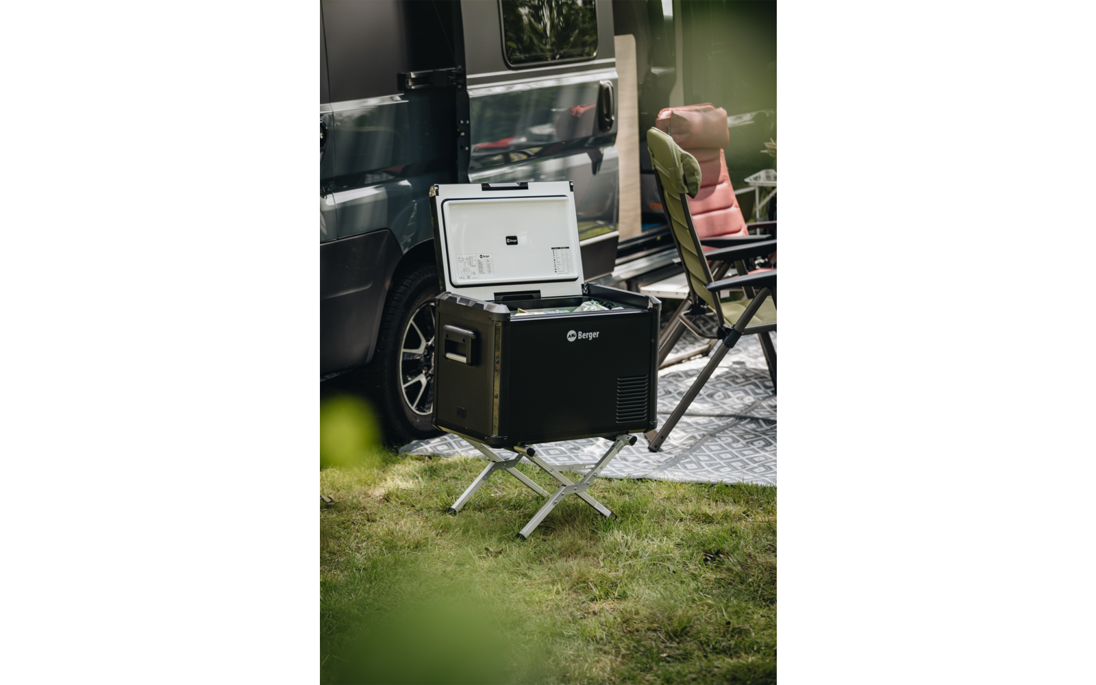 Offene schwarze tragbare Kühlbox auf Metallständer vor einem Van, daneben ein gepolsterter Campingstuhl und eine Bodenmatte auf Gras