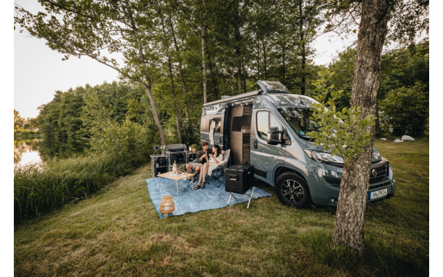 Grauer Camper-Van mit geöffneter Seitentür auf einer Wiese am See; drei Personen sitzen auf einer blauen Picknickdecke und Campingstühlen, Laterne und Lautsprecher daneben, Bäume im Hintergrund