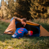 Person sitting at the entrance of an orange tent holding a blue sleeping bag; grass and trees in the background