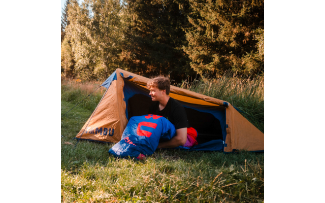 Person sitting at the entrance of an orange tent holding a blue sleeping bag; grass and trees in the background