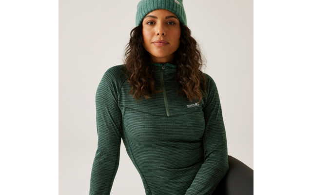 Femme aux longs cheveux bouclés portant un pull vert et un bonnet vert, assise devant un fond blanc