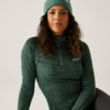 Frau mit langem lockigem Haar trägt grünen Pullover und grüne Mütze, sitzt vor weißem Hintergrund