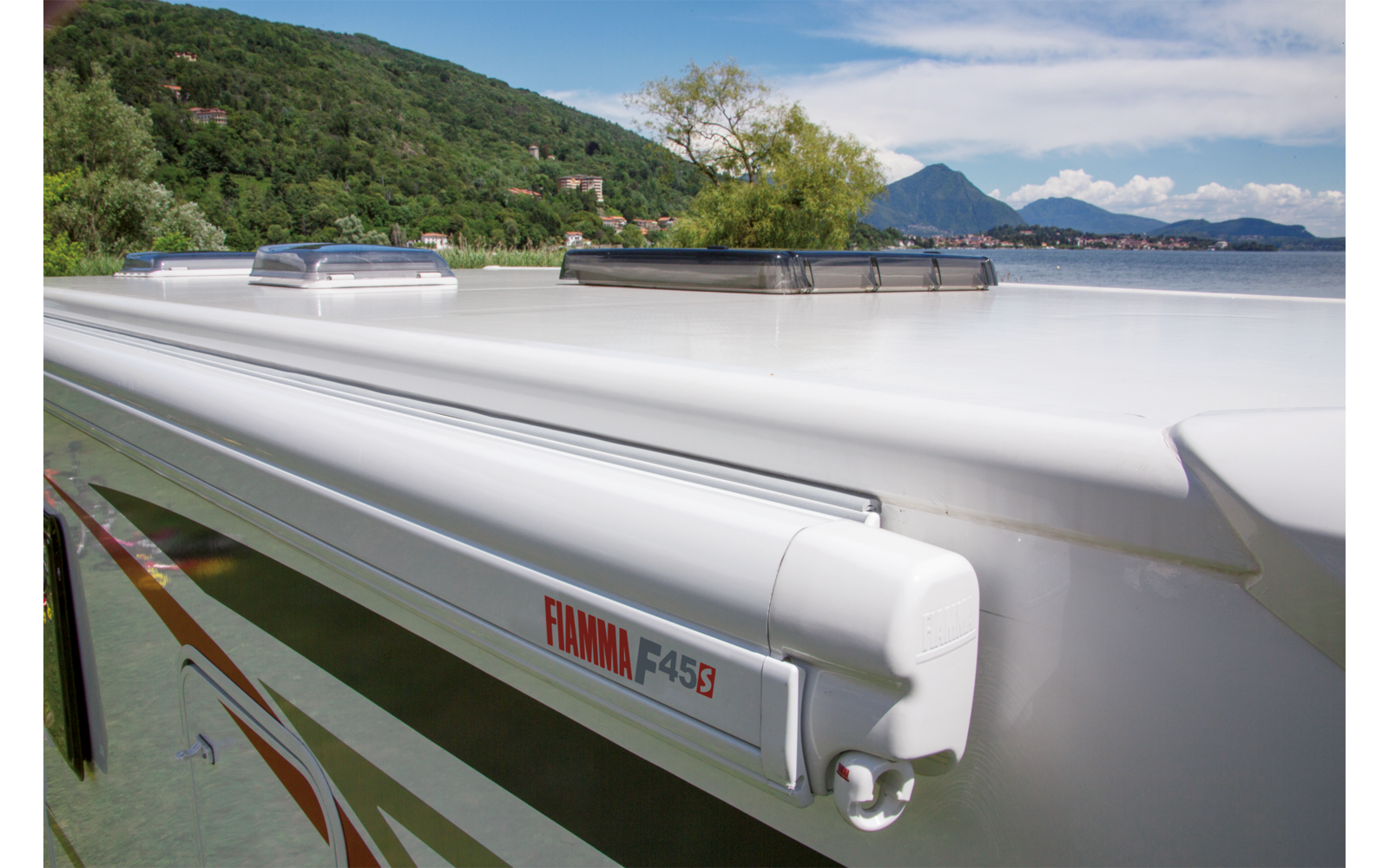 Dak van een witte camper met twee dakluiken en een opgerolde luifel met opschrift "Fiamma F45S", op de achtergrond een meer, bomen en bergen onder een blauwe lucht