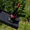 Rote Weinflasche mit rotem Schraubverschluss und Etikett 'METZGER ROT' in schwarzem Flaschenhalter auf schwarzem Tablett vor grünem Gartenhintergrund
