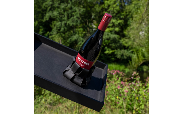 Rote Weinflasche mit rotem Schraubverschluss und Etikett 'METZGER ROT' in schwarzem Flaschenhalter auf schwarzem Tablett vor grünem Gartenhintergrund