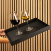 Hände halten schwarzes rechteckiges Tablett mit zwei Weingläsern (links Rotwein, rechts Weißwein) vor vertikaler Holzlattenwand