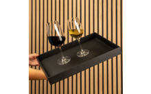 Hände halten schwarzes rechteckiges Tablett mit zwei Weingläsern (links Rotwein, rechts Weißwein) vor vertikaler Holzlattenwand