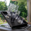 Schwarzes rechteckiges Tablett mit fünf Gläsern unterschiedlicher Form (zwei schlanke hohe Gläser, zwei mittelhohe Gläser, ein kurzes Glas) auf der Spülenkante; im Hintergrund Bäume, Wasser und rosa Blumen